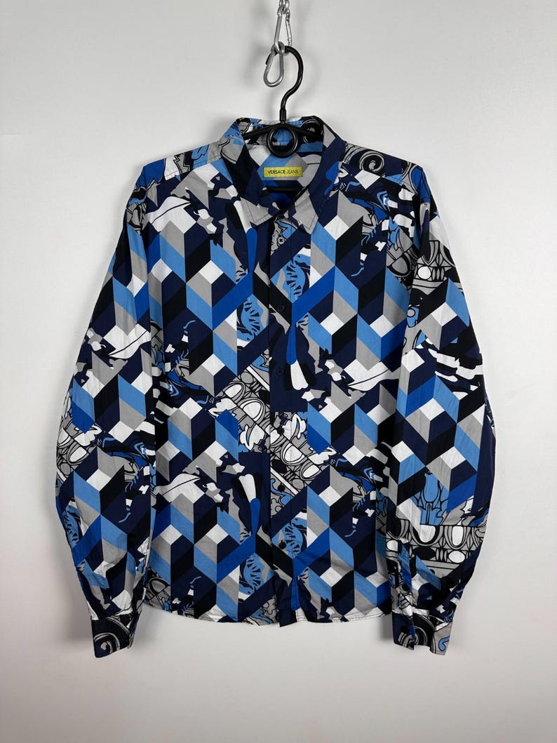 May include: A long-sleeved shirt with a geometric cube pattern in blue, grey, and black. The shirt also features a repeating design of white and grey decorative elements. The label reads "VERSACE JEANS".