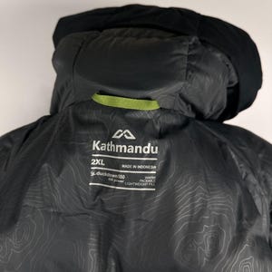 Puede incluir: Chaqueta con capucha Kathmandu 2XL negra. La chaqueta presenta una capucha negra y un detalle de lazo verde. El forro interior tiene un patr&oacute;n topogr&aacute;fico. La chaqueta tiene el logotipo de Kathmandu y el texto "duckdown 550 fill power" y "warm packable lightweight fill".