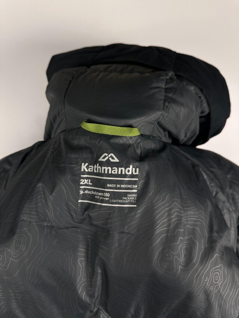 Puede incluir: Chaqueta con capucha Kathmandu 2XL negra. La chaqueta presenta una capucha negra y un detalle de lazo verde. El forro interior tiene un patr&oacute;n topogr&aacute;fico. La chaqueta tiene el logotipo de Kathmandu y el texto "duckdown 550 fill power" y "warm packable lightweight fill".