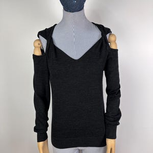 May include: Dark gray long-sleeve sweater with a V-neck and open shoulder design. The sweater is displayed on a mannequin with a blue and white striped shirt underneath. The mannequin also has a dark blue hat.