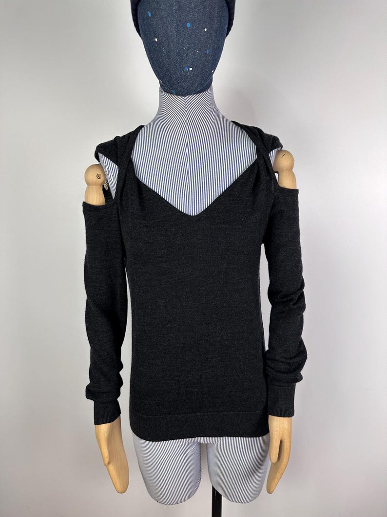 May include: Dark gray long-sleeve sweater with a V-neck and open shoulder design. The sweater is displayed on a mannequin with a blue and white striped shirt underneath. The mannequin also has a dark blue hat.
