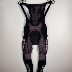Puede incluir: Culote de ciclismo negro y morado con el nombre de la marca "X BIONIC" impreso en las piernas. El culote está colgado de una percha negra sobre un fondo blanco. El culote tiene una badana acolchada y tirantes.
