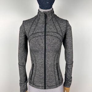 May include: A gray athletic jacket with a full-length black zipper and two front pockets. The jacket has a high collar and long sleeves with thumbholes. The fabric has a textured appearance. The jacket is displayed on a mannequin.