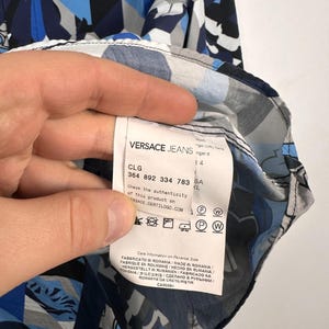 May include: A Versace Jeans shirt featuring a geometric and floral design in blue, grey, and white. The label is visible, displaying the brand name and product details.