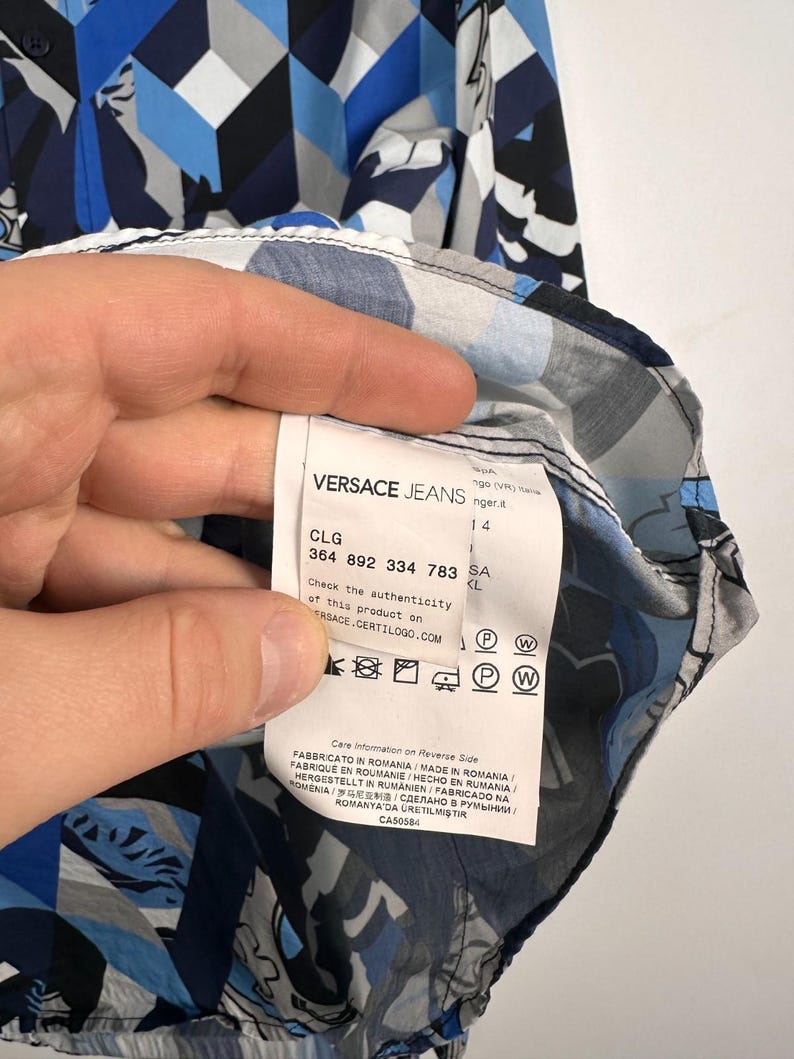 May include: A Versace Jeans shirt featuring a geometric and floral design in blue, grey, and white. The label is visible, displaying the brand name and product details.