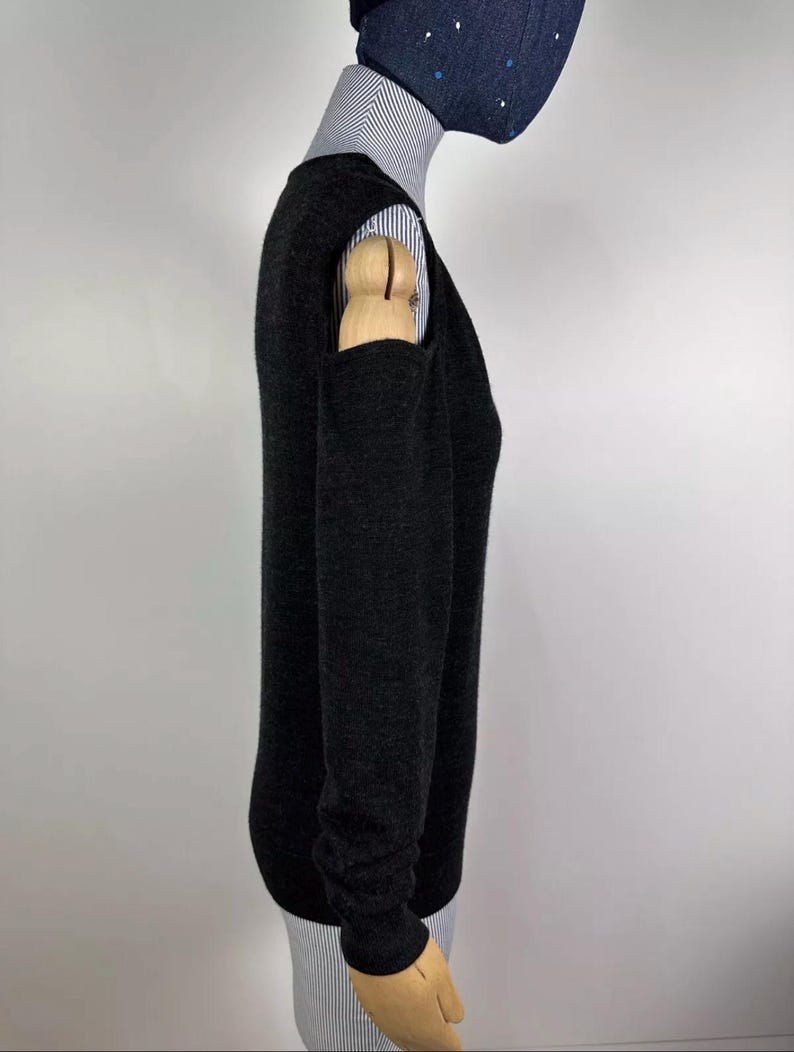 May include: A dark gray sweater with long sleeves and a crew neck. The sweater has a unique design with cut-out details on the shoulders. The sweater is worn over a striped collared shirt. The mannequin is wearing a dark blue face mask with white dots.