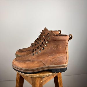 Könnte beinhalten: Braune Lederstiefel mit Mokassin-Zehen und Metallösen. Die Stiefel haben ein abgenutztes Aussehen und stehen auf einem Holzschemel. Das Timberland-Logo ist sichtbar. Die Stiefel sind ca. 20 cm hoch.