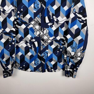 May include: A long-sleeved shirt with a striking geometric design in blue, grey, and white. The pattern incorporates cube shapes and floral motifs. The shirt has a traditional collar and buttoned cuffs.