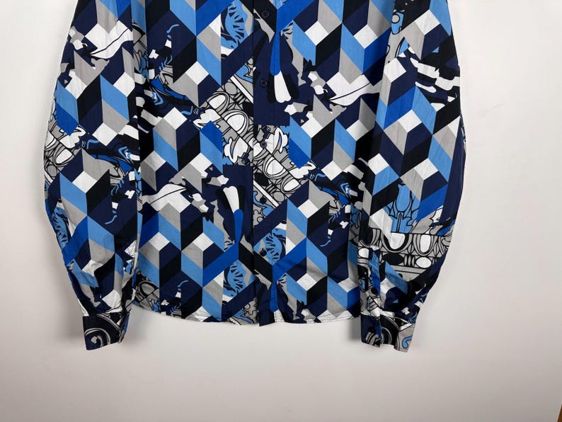 May include: A long-sleeved shirt with a striking geometric design in blue, grey, and white. The pattern incorporates cube shapes and floral motifs. The shirt has a traditional collar and buttoned cuffs.