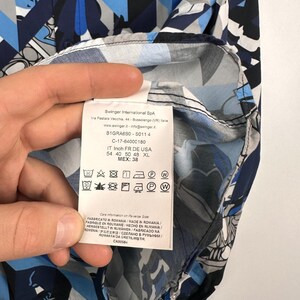 May include: A patterned button-down shirt in blue, white, and grey geometric shapes. The label shows care instructions and sizing details. The shirt is made in Romania. Measurements are in centimeters.