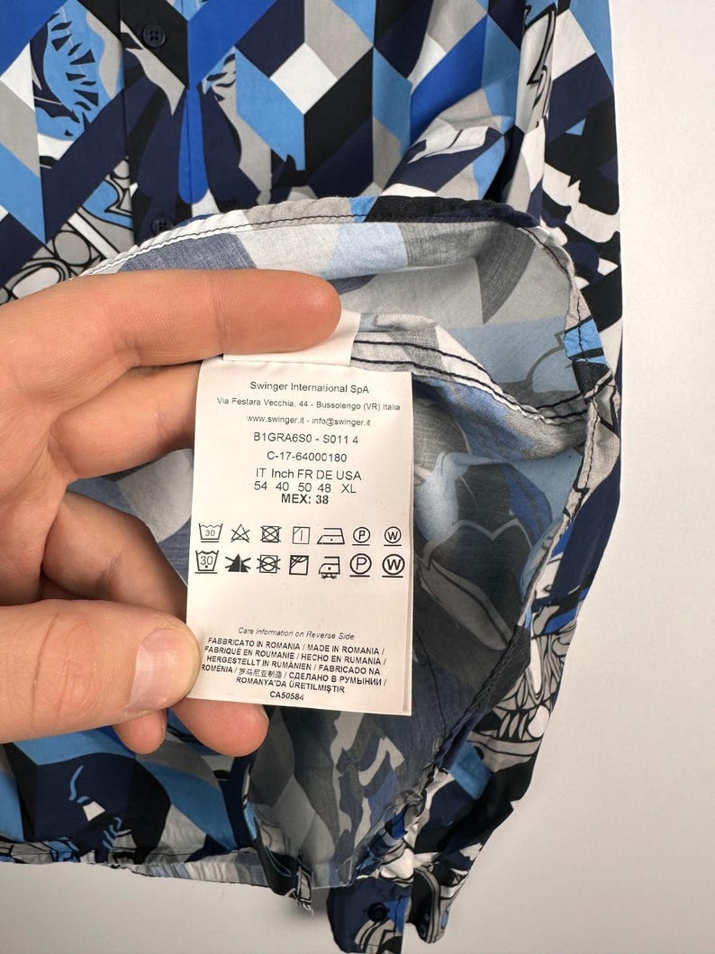 May include: A patterned button-down shirt in blue, white, and grey geometric shapes. The label shows care instructions and sizing details. The shirt is made in Romania. Measurements are in centimeters.