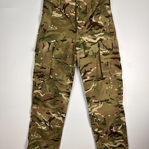 Könnte beinhalten: Tarnfarbene Cargohose mit mehreren Taschen und einem Knopfverschluss. Die Hose hat eine Mischung aus grünen, braunen und beigen Farben in einem militärischen Muster. Die Hose hängt an einem Kleiderbügel.