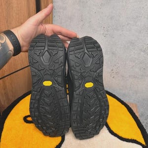 May include: Pair of black hiking boots with textured soles, featuring yellow Vibram logos. The soles have a rugged tread pattern for grip. The boots are displayed against a neutral background.