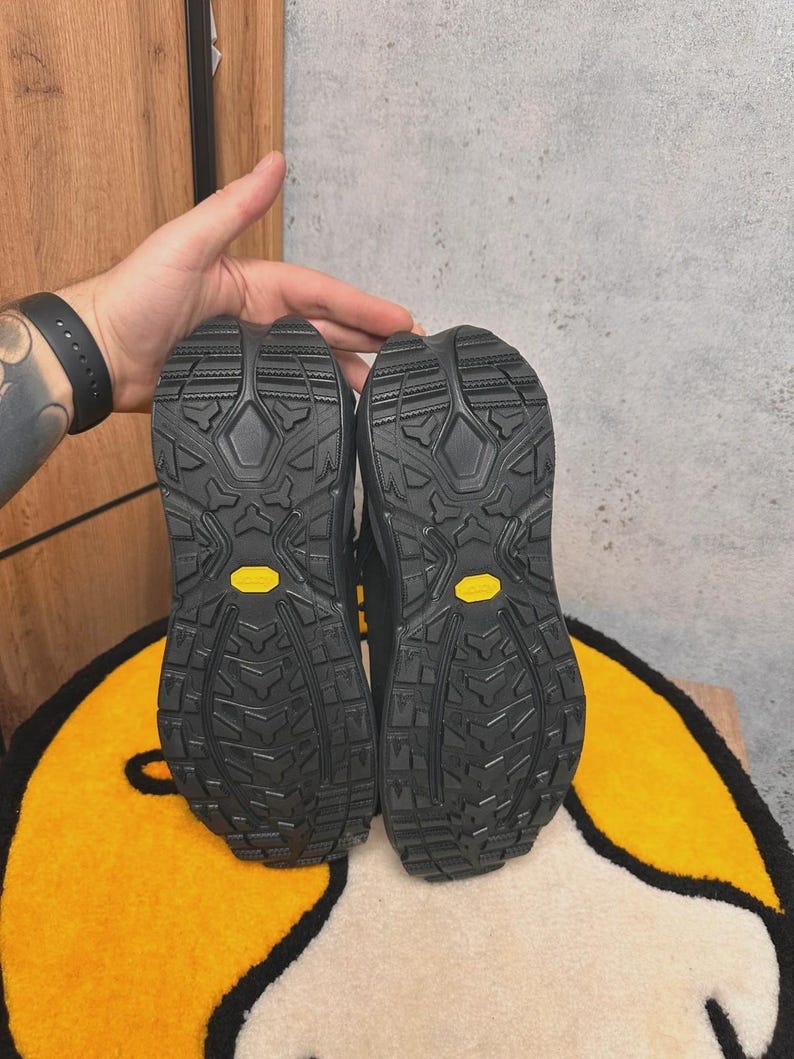 May include: Pair of black hiking boots with textured soles, featuring yellow Vibram logos. The soles have a rugged tread pattern for grip. The boots are displayed against a neutral background.