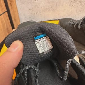 May include: Close-up of a black athletic shoe with gray laces. The shoe's interior reveals a label with the brand name "HOKA ONE ONE" and size information: US 12D (11.5 46⅔) 30. The shoe is designed for running or other athletic activities.