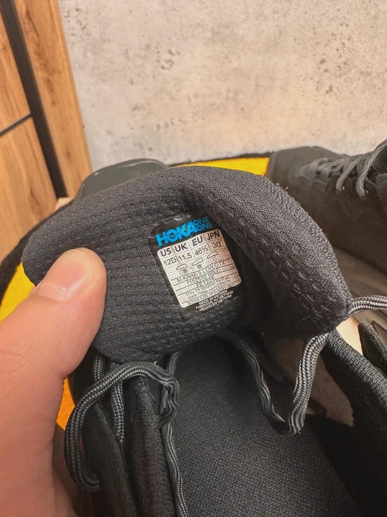 May include: Close-up of a black athletic shoe with gray laces. The shoe's interior reveals a label with the brand name "HOKA ONE ONE" and size information: US 12D (11.5 46⅔) 30. The shoe is designed for running or other athletic activities.