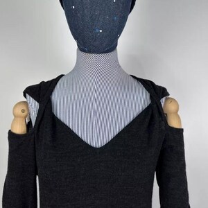 May include: A dark gray, long-sleeved sweater with a V-neck and cut-out shoulder design. The sweater is displayed on a mannequin with a striped shirt underneath and a dark blue beanie with white speckles.