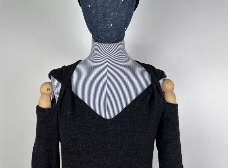 May include: A dark gray, long-sleeved sweater with a V-neck and cut-out shoulder design. The sweater is displayed on a mannequin with a striped shirt underneath and a dark blue beanie with white speckles.