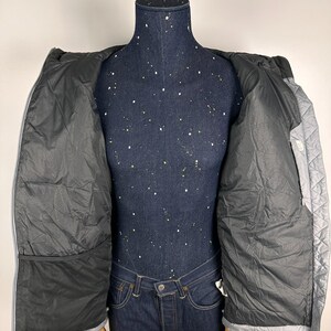 Puede incluir: Chaqueta acolchada gris claro con interior negro, expuesta abierta sobre un maniqu&iacute;. La chaqueta tiene capucha y cierre de cremallera. El interior tiene un bolsillo grande a cada lado. El maniqu&iacute; lleva vaqueros azules.