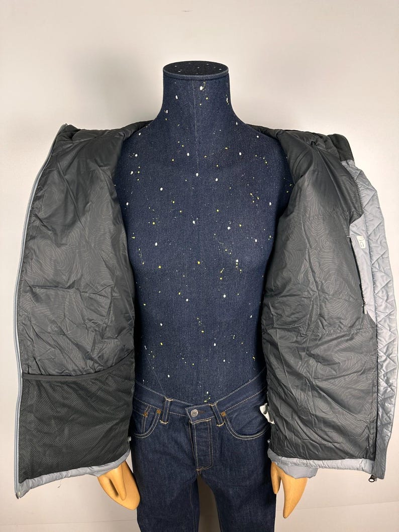 Puede incluir: Chaqueta acolchada gris claro con interior negro, expuesta abierta sobre un maniqu&iacute;. La chaqueta tiene capucha y cierre de cremallera. El interior tiene un bolsillo grande a cada lado. El maniqu&iacute; lleva vaqueros azules.