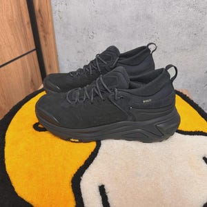 May include: Pair of black athletic shoes with thick soles and laces, resting on a yellow and white rug. The shoes have a textured design and a pull tab at the heel. The word "Vibram" is visible on the sole.