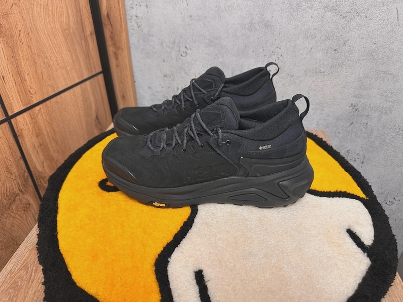 May include: Pair of black athletic shoes with thick soles and laces, resting on a yellow and white rug. The shoes have a textured design and a pull tab at the heel. The word "Vibram" is visible on the sole.