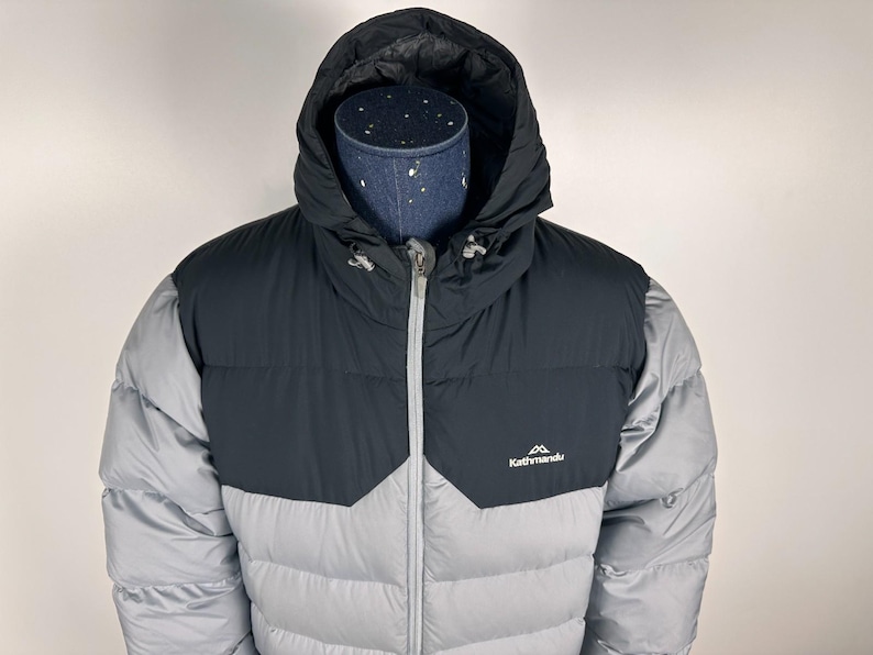 Puede incluir: Chaqueta acolchada con capucha gris y negra, con cremallera completa y el logotipo "Kathmandu". La chaqueta tiene un dise&ntilde;o acolchado y un cuello alto para mayor calidez.
