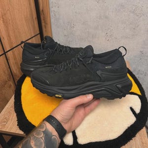 May include: Pair of black Hoka hiking shoes with Vibram soles. The shoes have a Gore-Tex label and are laced up. The shoes are on a yellow and cream-colored rug.