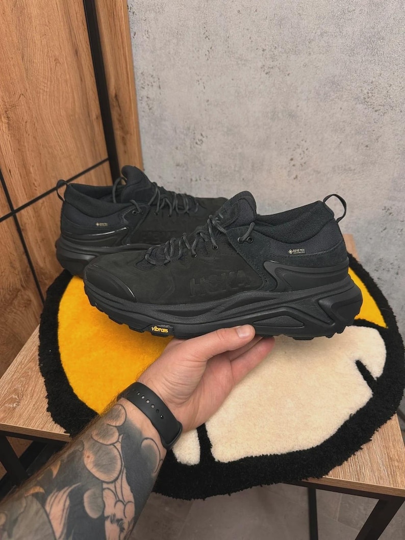 May include: Pair of black Hoka hiking shoes with Vibram soles. The shoes have a Gore-Tex label and are laced up. The shoes are on a yellow and cream-colored rug.