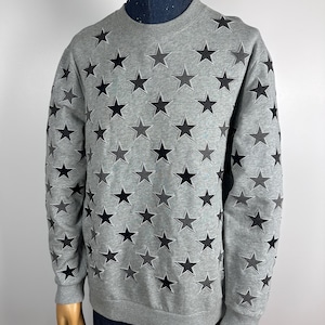 Pode incluir: Uma camisa de moletom cinza com um padrão de estrelas pretas. As estrelas são de tamanhos variados e estão dispostas em um padrão aleatório.