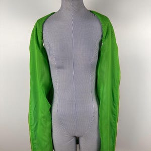 May include: A bright green jacket with long sleeves is draped over a mannequin. The jacket has a textured pattern and gold-colored zippers. The mannequin is striped in white and blue, and wears a dark blue cap.