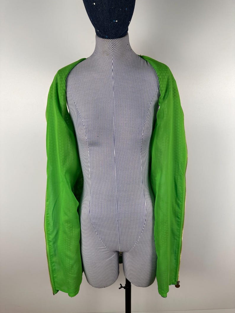 May include: A bright green jacket with long sleeves is draped over a mannequin. The jacket has a textured pattern and gold-colored zippers. The mannequin is striped in white and blue, and wears a dark blue cap.