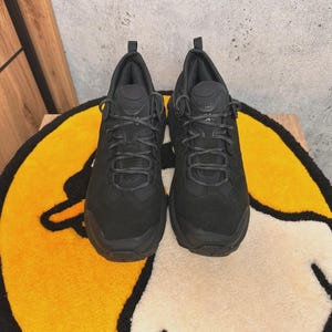 May include: A pair of all-black athletic shoes with a textured upper and a lace-up closure. The shoes are displayed on a yellow and white rug with a cartoon design. The shoes have a thick sole and a padded collar.