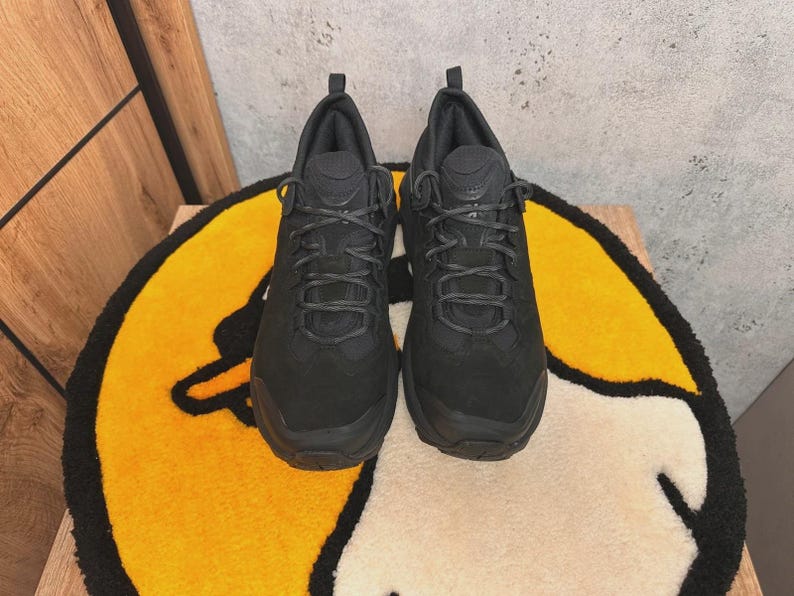 May include: A pair of all-black athletic shoes with a textured upper and a lace-up closure. The shoes are displayed on a yellow and white rug with a cartoon design. The shoes have a thick sole and a padded collar.