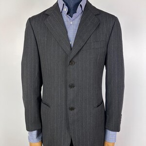 Pode incluir: Um blazer cinza carvão com riscas finas, com três botões pretos e lapelas entalhadas. O blazer é exibido em um manequim com uma camisa xadrez azul e branca por baixo. O blazer tem um corte clássico e sob medida.