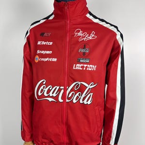 Può includere: Giacca a vento rossa con strisce bianche e nere sulle maniche. La giacca ha una chiusura a zip e un colletto alto. La parte anteriore della giacca presenta un grande logo Coca-Cola, un logo Coca-Cola più piccolo e il testo "LACTION". Ci sono anche diversi loghi di sponsor, tra cui "ACDetco", "Snapan" e "coopFrEAR".