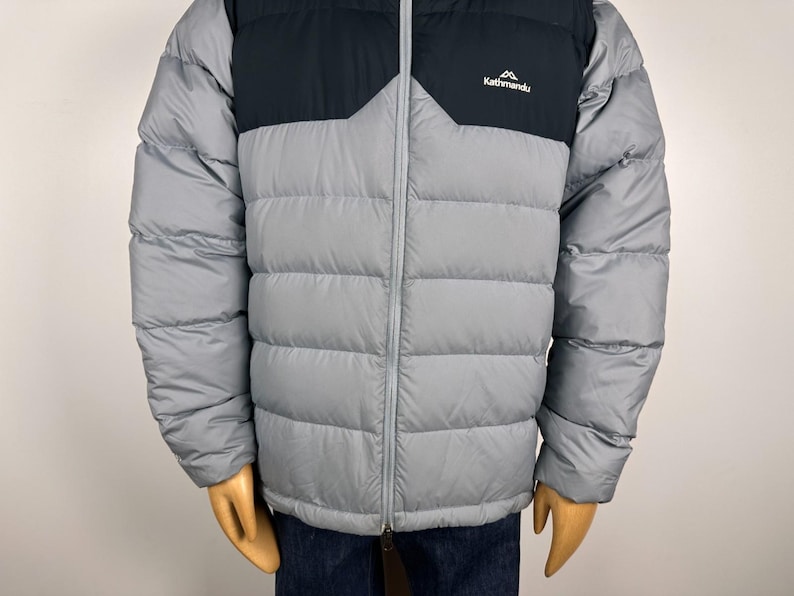 Puede incluir: Chaqueta acolchada gris y negra con cremallera completa. La chaqueta tiene una secci&oacute;n superior negra y una secci&oacute;n inferior gris. El logotipo de Kathmandu est&aacute; en la parte superior izquierda del pecho. Para clima fr&iacute;o.