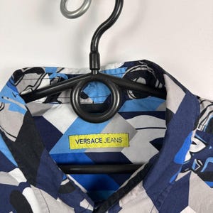 May include: A patterned shirt in blue, grey, and white geometric shapes. The shirt has a collar and a yellow label with "VERSACE JEANS". It is displayed on a black hanger. The shirt is a stylish piece of clothing.