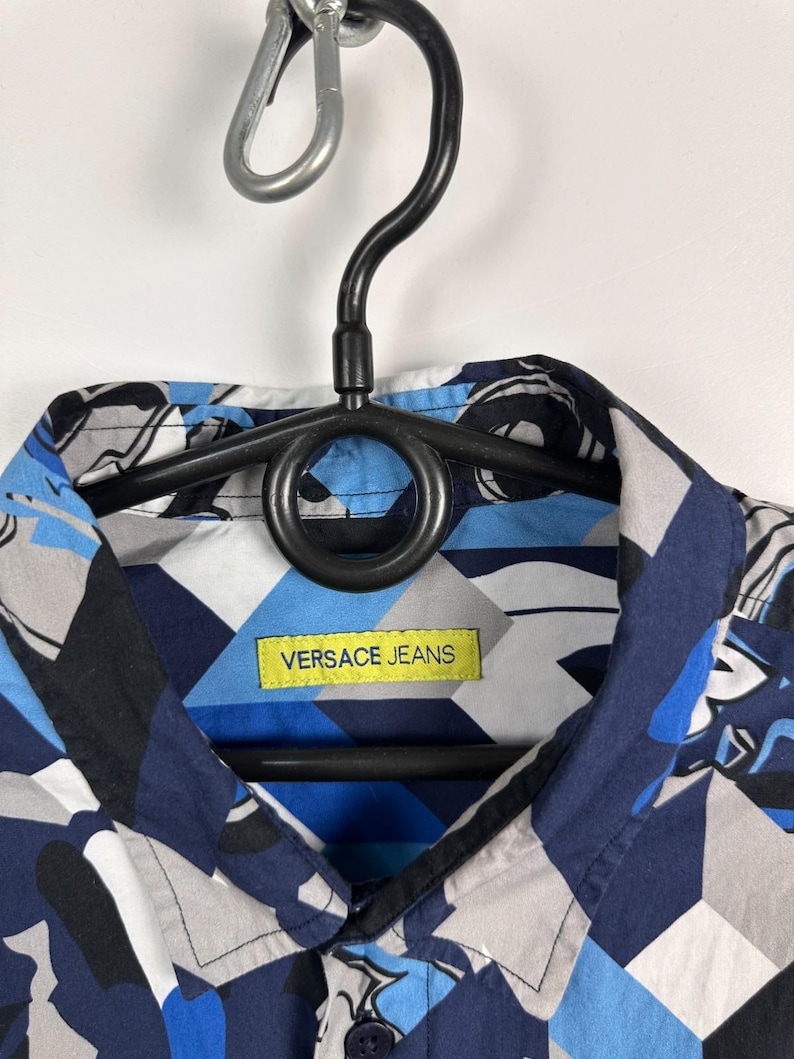 May include: A patterned shirt in blue, grey, and white geometric shapes. The shirt has a collar and a yellow label with "VERSACE JEANS". It is displayed on a black hanger. The shirt is a stylish piece of clothing.