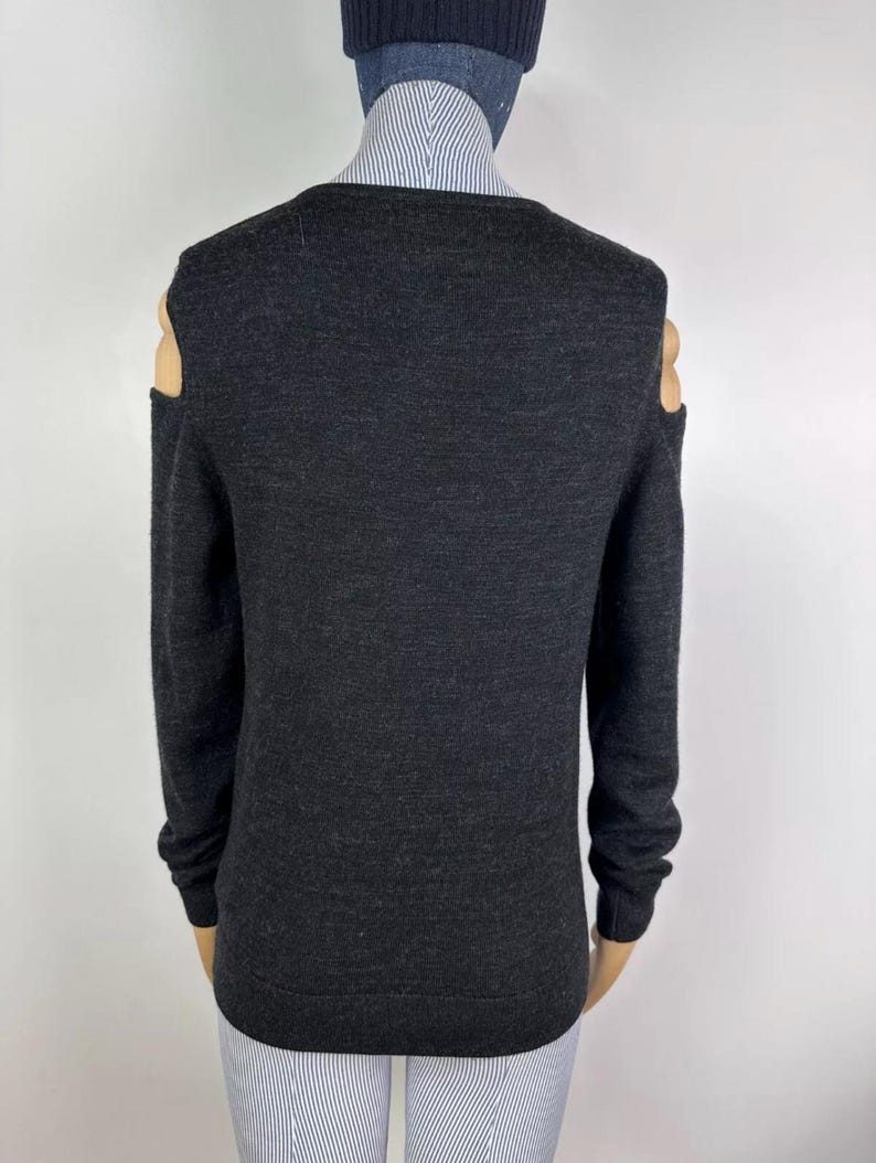 May include: Dark gray sweater with long sleeves and cut-out shoulder details. The sweater has a v-neck and is made of a soft, knitted material. The cut-out shoulder design adds a modern touch to the classic sweater style.