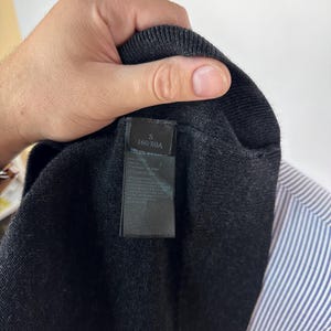 May include: Close-up of a dark gray knit sweater with a black label. The label reads "S 160/80A". A blue and white striped shirt is visible in the background.