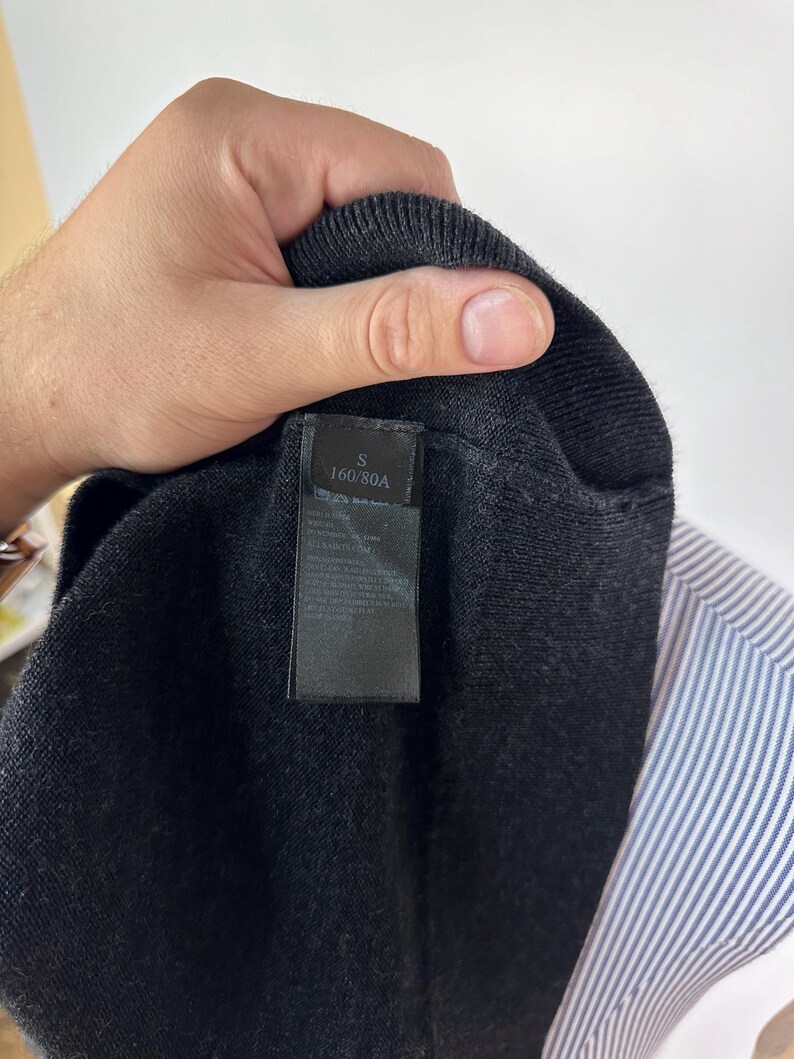 May include: Close-up of a dark gray knit sweater with a black label. The label reads "S 160/80A". A blue and white striped shirt is visible in the background.