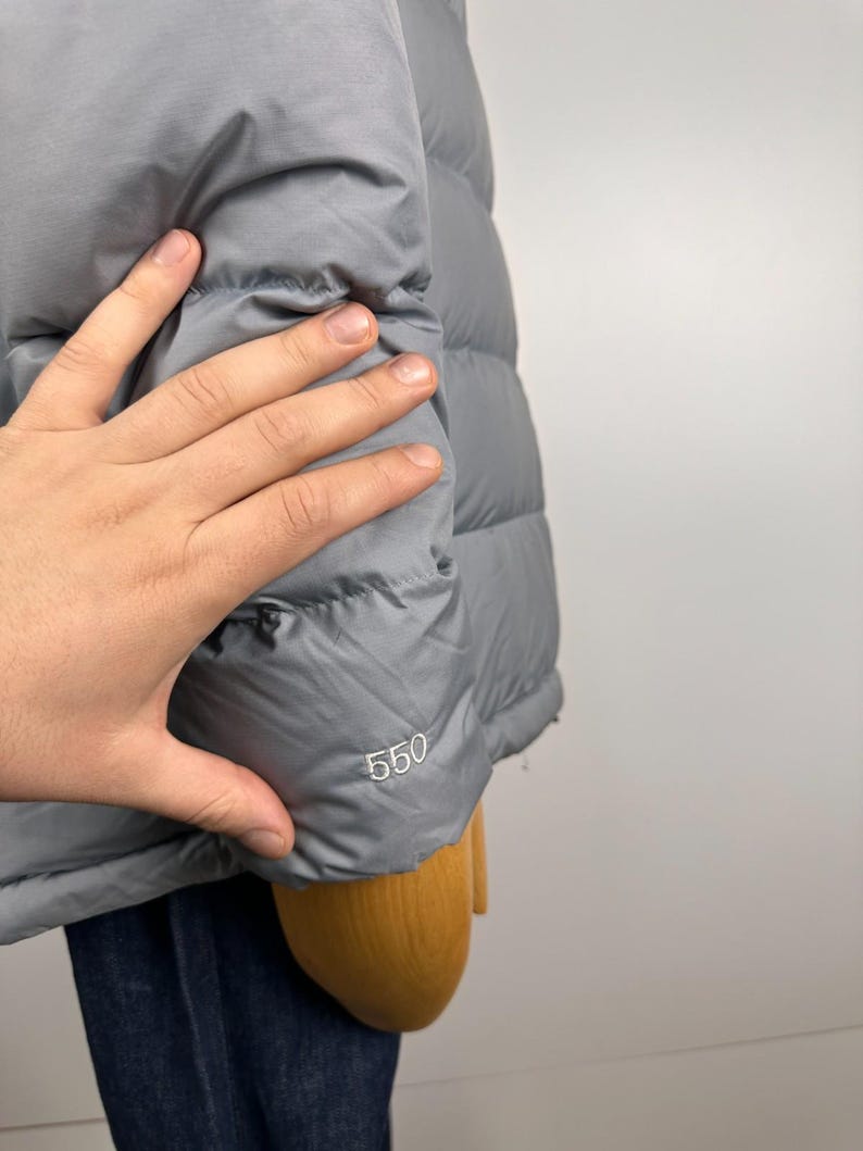 Puede incluir: Una chaqueta acolchada gris con un dise&ntilde;o acolchado. El n&uacute;mero "550" est&aacute; impreso en la manga. La chaqueta est&aacute; siendo sostenida por una persona.