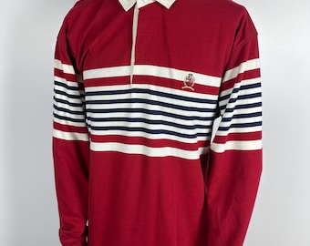 May include: A red long-sleeved rugby shirt with white and navy blue horizontal stripes on the sleeves. It features a white collar and a small emblem on the chest. The shirt is displayed on a mannequin.