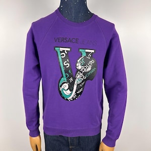 Puede incluir: Una sudadera morada con el texto "VERSACE JEANS" en el pecho. El gráfico en la parte delantera presenta una "V" estilizada con detalles en negro, blanco y verde azulado. La sudadera se muestra en un maniquí.
