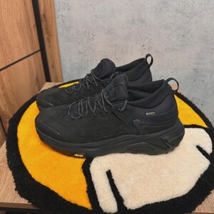 May include: A pair of black athletic shoes with dark laces and a textured upper. The shoes are on a yellow, black, and white patterned rug. The shoes have a Gore-Tex label.