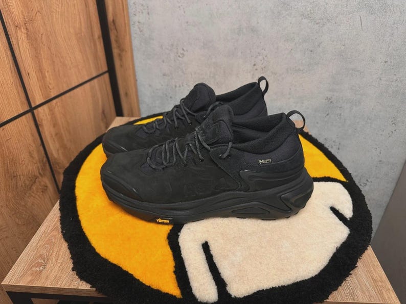 May include: A pair of black athletic shoes with dark laces and a textured upper. The shoes are on a yellow, black, and white patterned rug. The shoes have a Gore-Tex label.