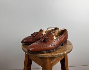 Puede incluir: Un par de mocasines con borlas de cuero marrón con perforaciones decorativas. Los zapatos se exhiben en un pequeño taburete de madera, destacando su diseño clásico y artesanía.