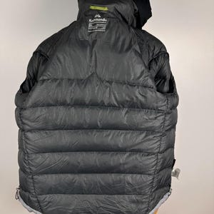 Puede incluir: Chaqueta acolchada negra con capucha. La chaqueta presenta un dise&ntilde;o acolchado horizontal y un ribete gris en el bajo. El logotipo de Kathmandu es visible en la parte trasera de la capucha. La chaqueta es talla XXL.
