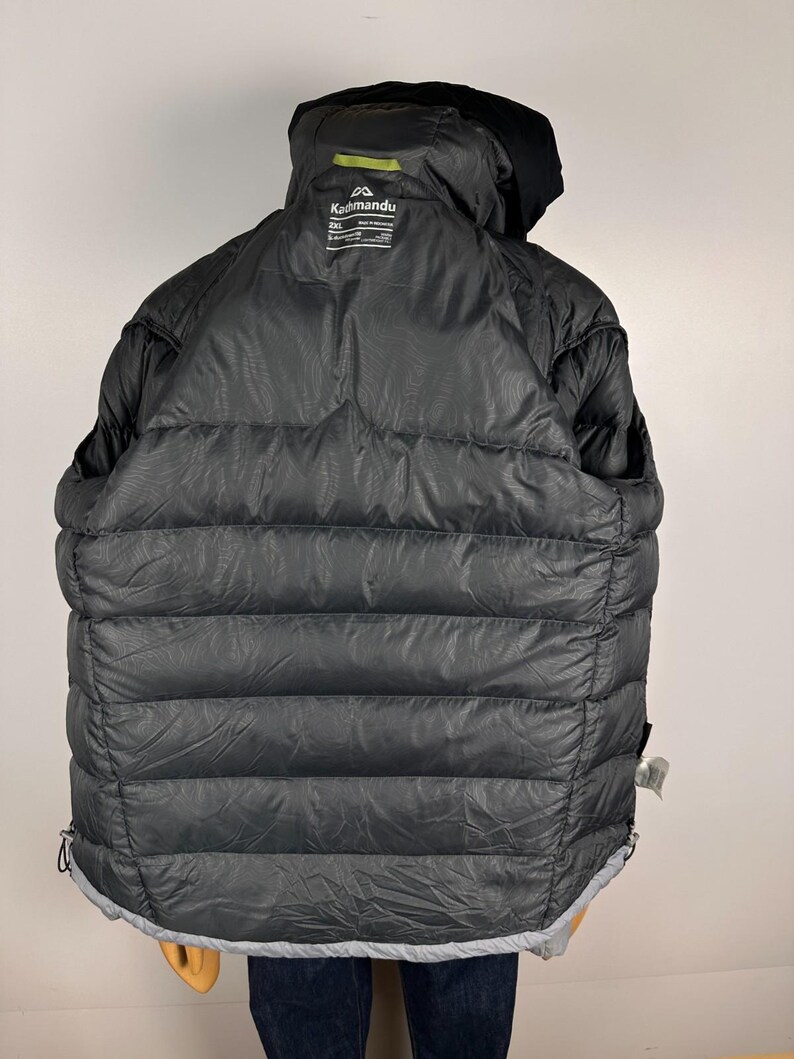 Puede incluir: Chaqueta acolchada negra con capucha. La chaqueta presenta un dise&ntilde;o acolchado horizontal y un ribete gris en el bajo. El logotipo de Kathmandu es visible en la parte trasera de la capucha. La chaqueta es talla XXL.